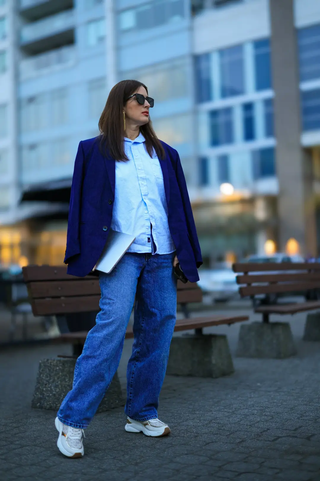 A woman in a blazer and jeans walks across a paved plaza while carrying a laptop, city buildings blurred behind her.