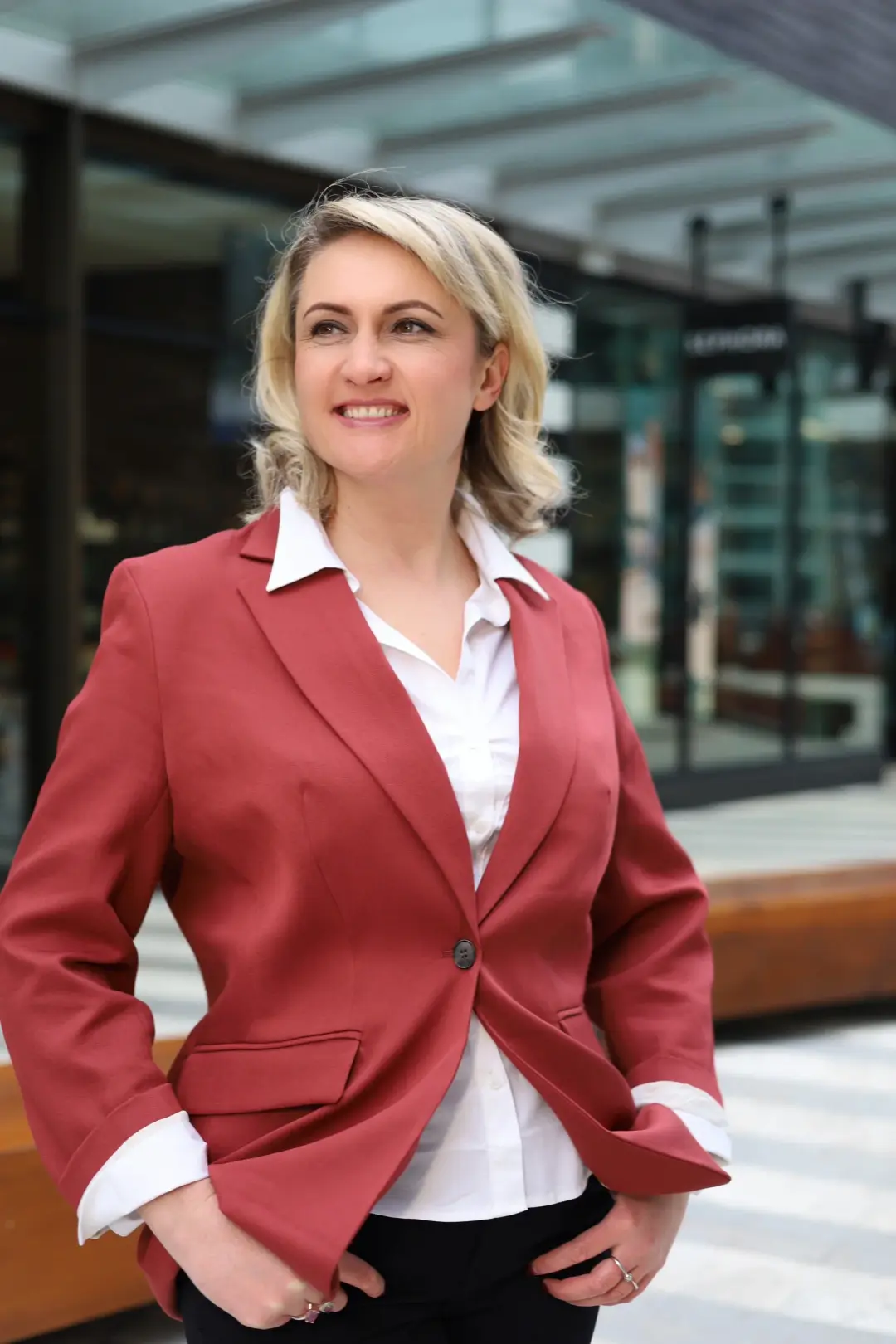 A smiling woman in a rust-colored blazer stands with her hands in her pockets outside a glass storefront.