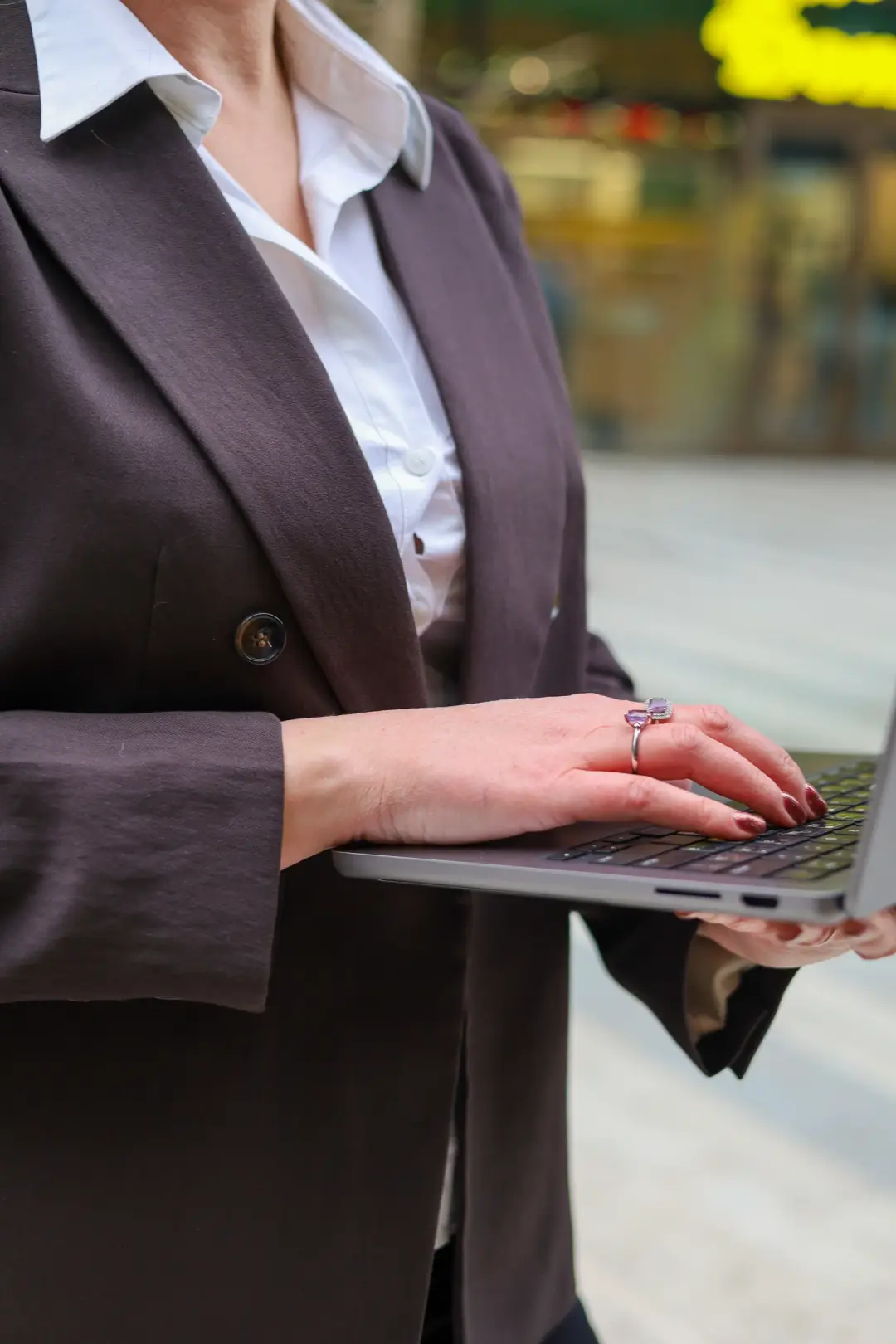 A close-up of a hand typing on a laptop keyboard, dressed in a tailored blazer and white shirt with a blurred street scene beyond.