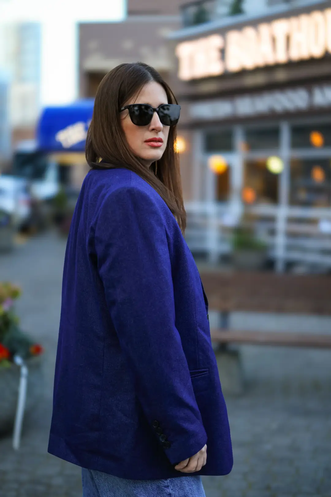 A woman in a purple blazer and sunglasses turns her head in profile on a street corner, with an illuminated restaurant sign glowing behind her.
