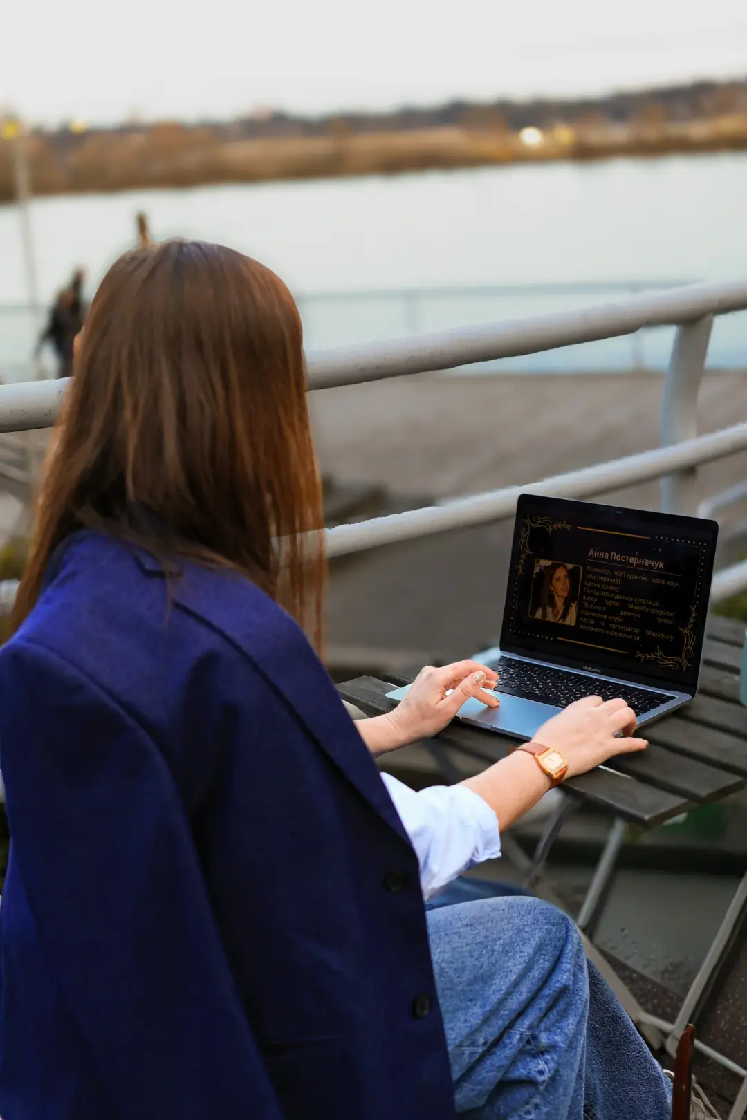 Seen from behind, a woman with long auburn hair works on a laptop at an outdoor table beside a waterfront railing.