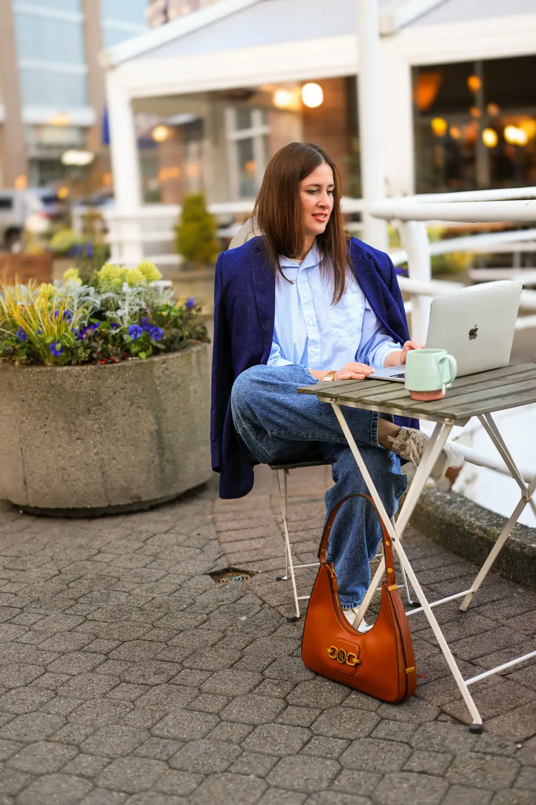 A woman sits cross-legged at a small bistro table with a laptop and mug, a leather bag resting on the ground beside her.