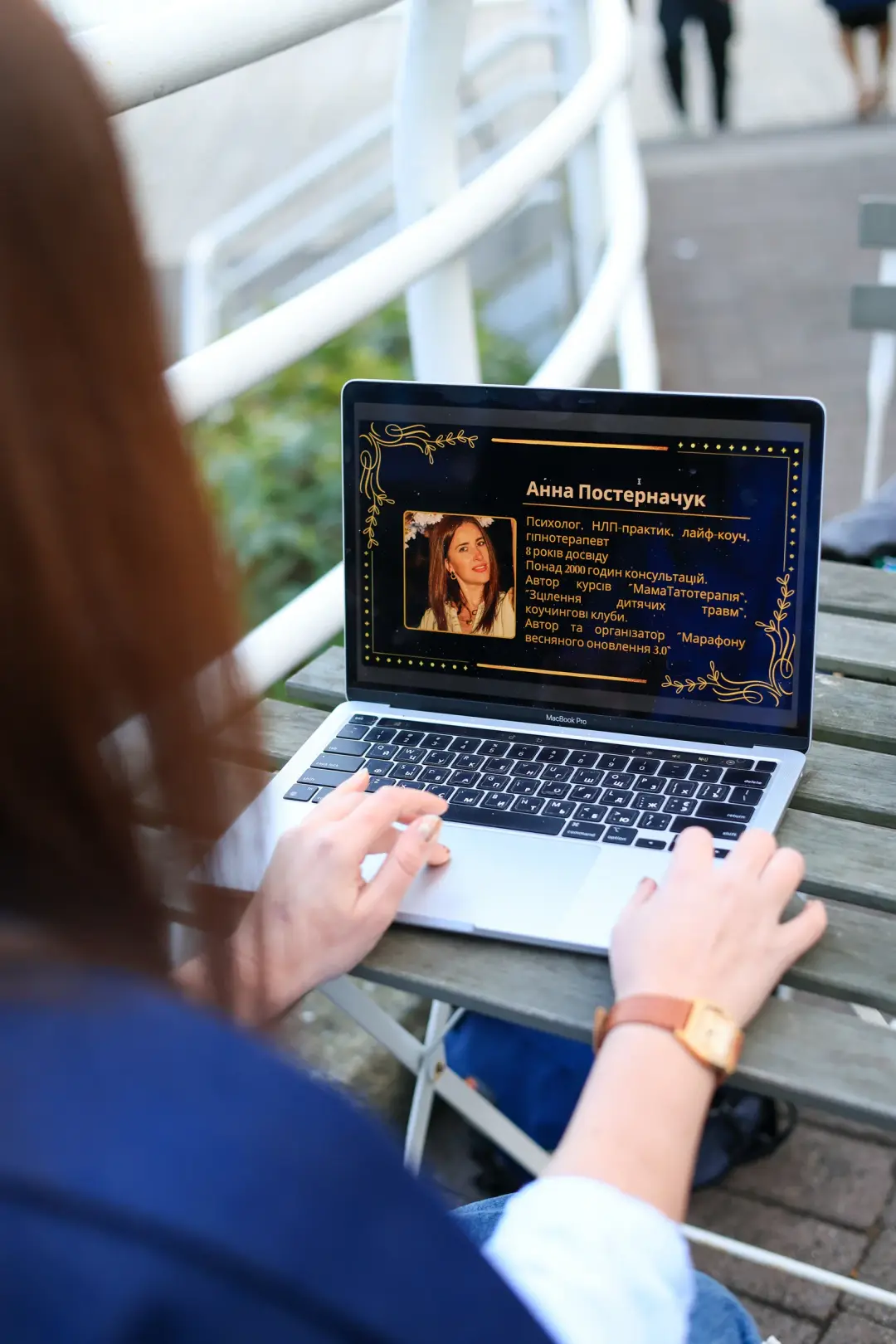 An over-the-shoulder view of hands typing on a laptop displaying a presentation slide, outdoors on a wooden table.