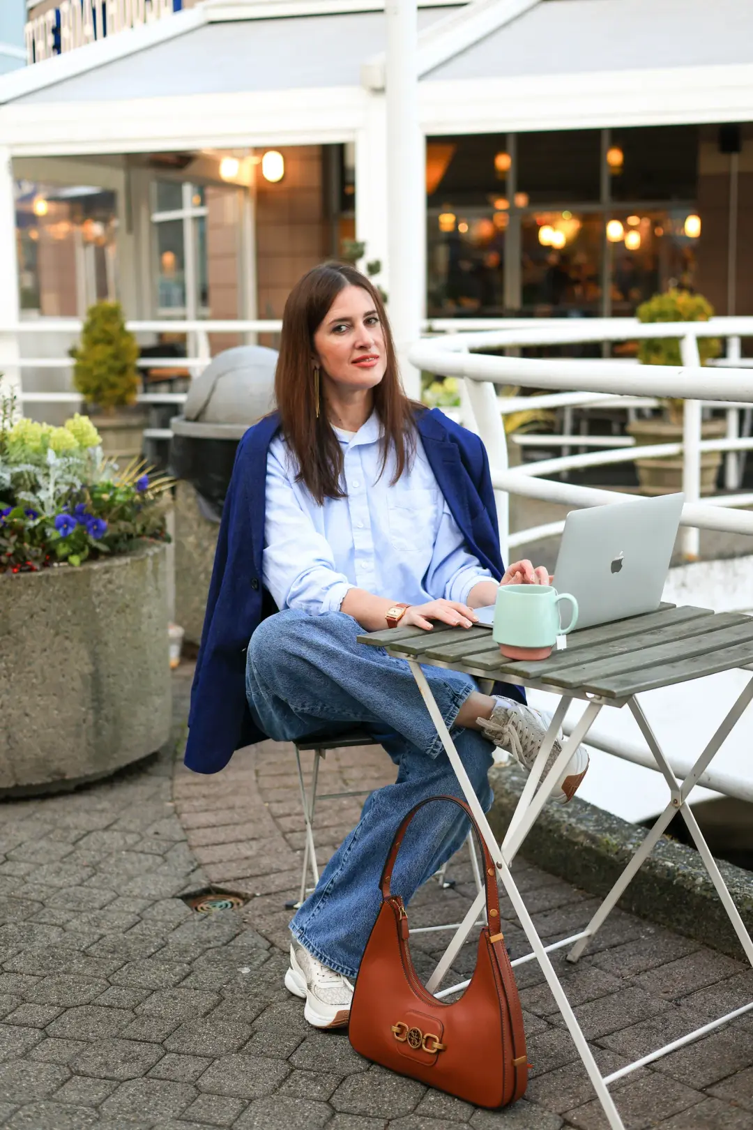 A woman in a light blue shirt and blazer sits at an outdoor cafe table with a laptop and mug, looking directly at the camera.