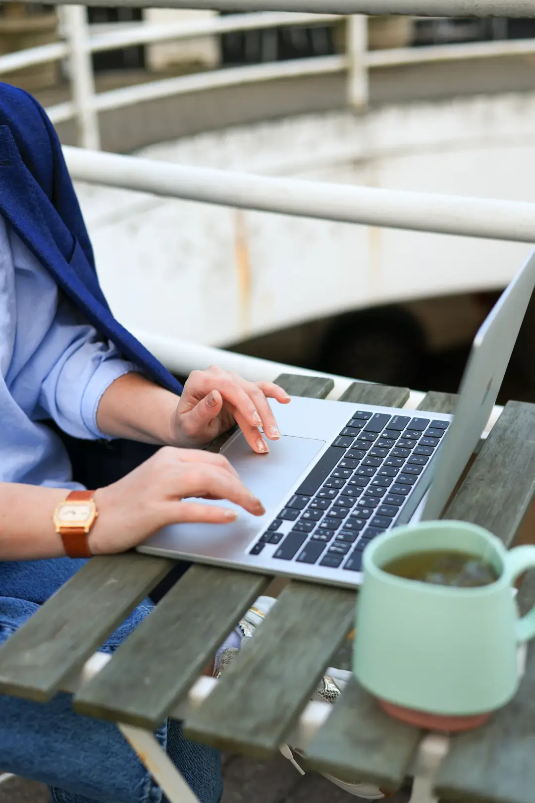 A close-up of hands typing on a laptop trackpad and keyboard, with a mug and blurred railing visible on an outdoor table.
