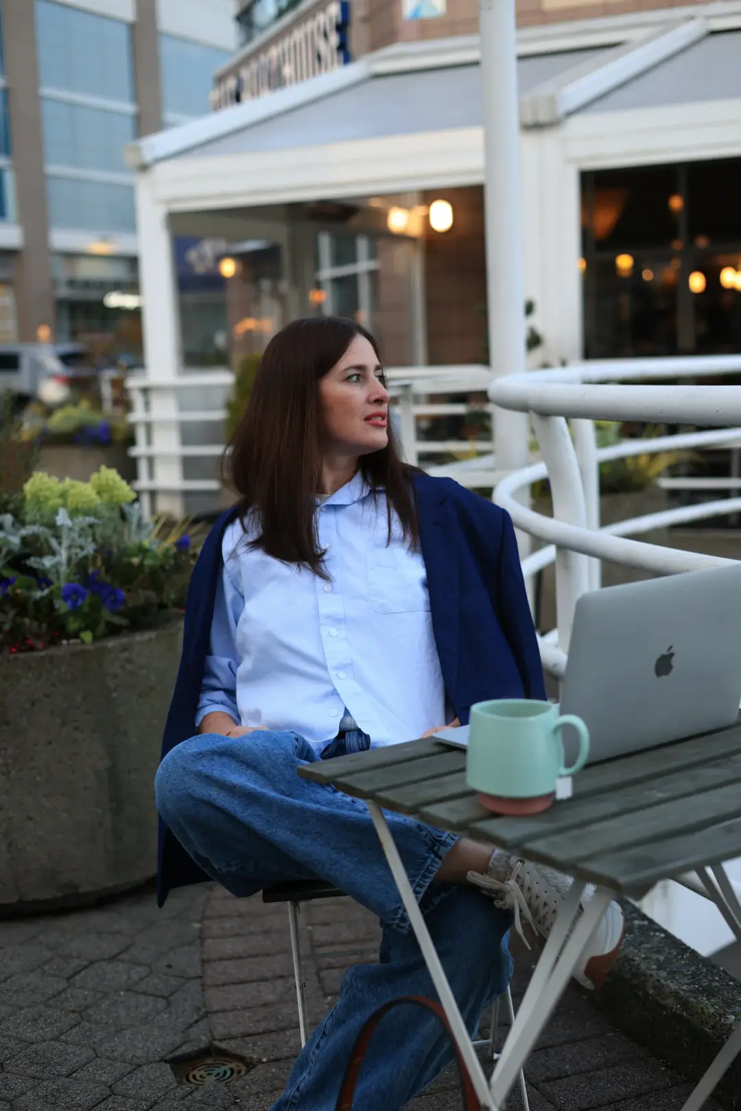 A woman in a blazer sits at an outdoor table with a laptop and mug, looking off to the side in conversation.