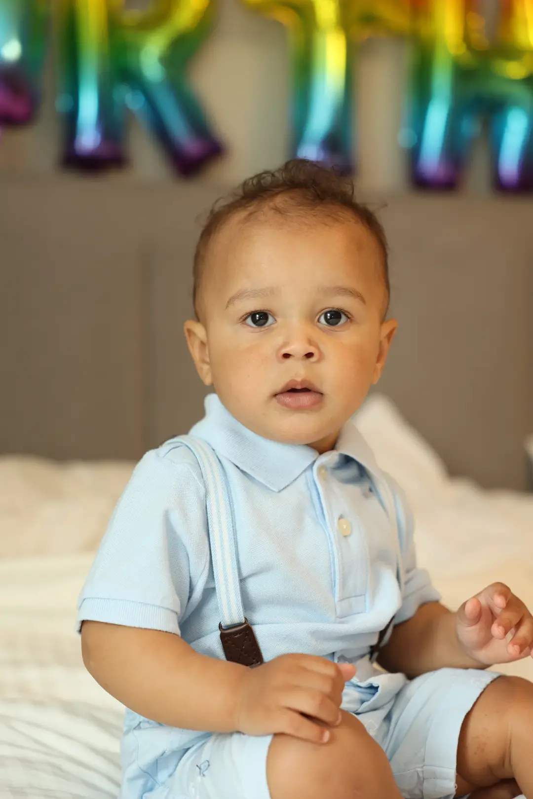 A toddler boy in a light blue romper with suspenders sits on a bed in front of blurred rainbow balloon letters.