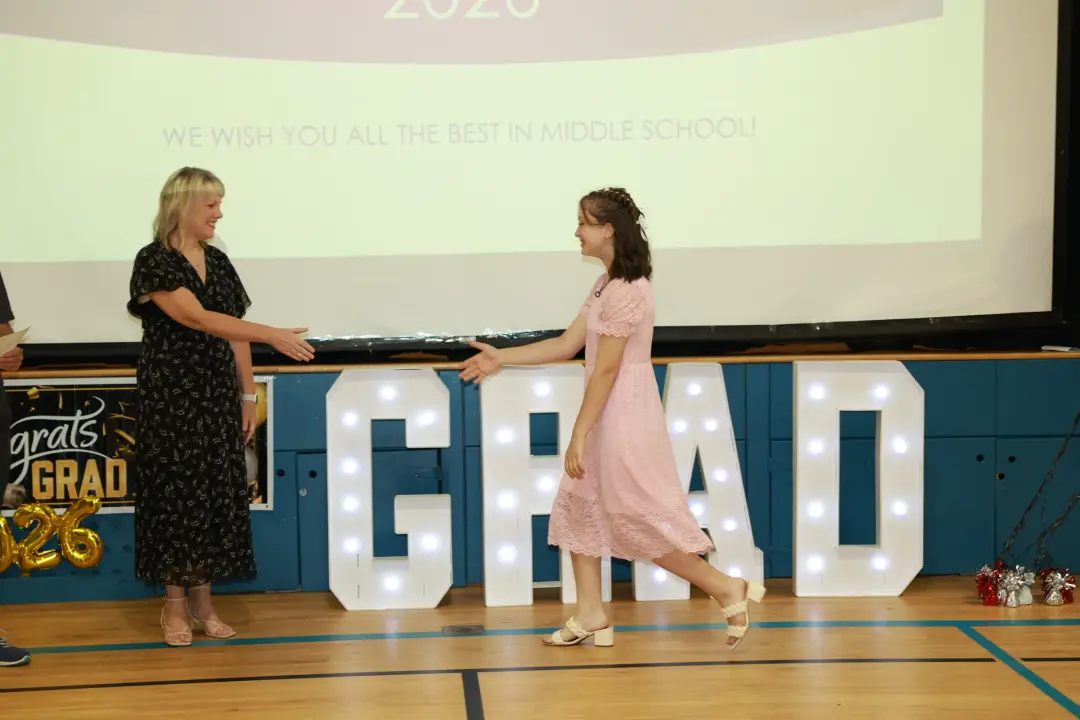A graduate in a pink lace dress crosses the gym floor to shake hands beside glowing marquee letters spelling GRAD.