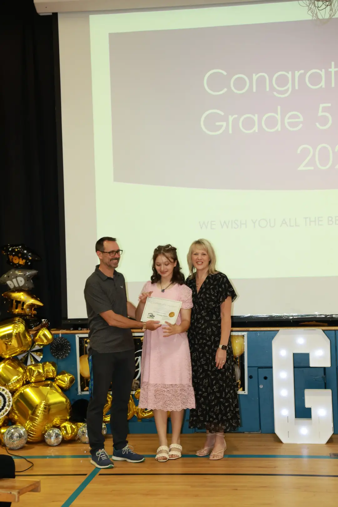 A young graduate poses with two adults on a decorated stage, holding a certificate amid gold balloon clusters.