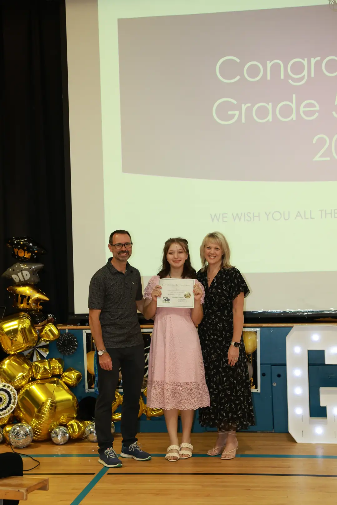 Two adults celebrate with a certificate-holding graduate on a festive stage framed by gold balloons.
