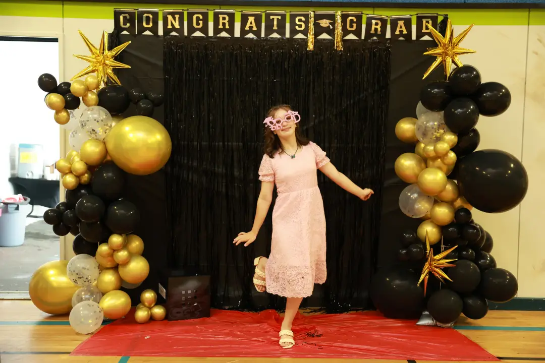 A graduate poses playfully in novelty grad glasses under a gold and black balloon arch on a red carpet.
