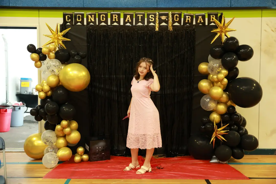 A graduate pauses to touch her hair beneath a gold and black balloon arch on a red carpet.