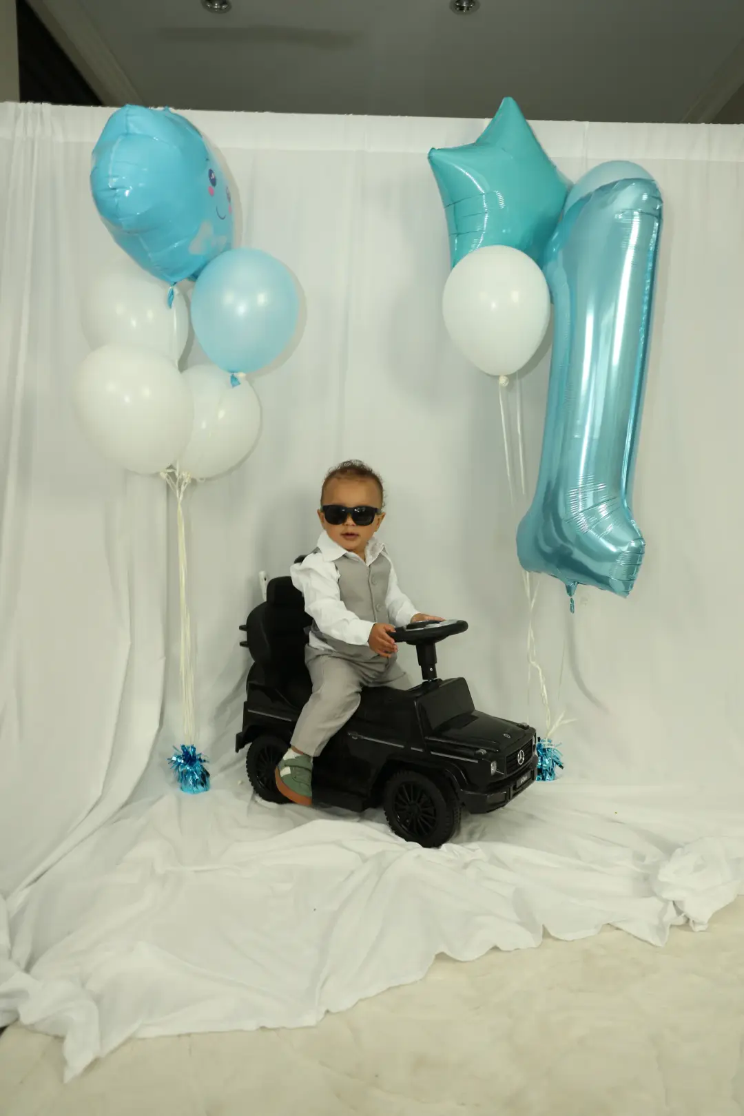 A toddler in sunglasses and a grey vest sits on a black toy ride-on car beside blue and white balloons and a large number-one balloon.