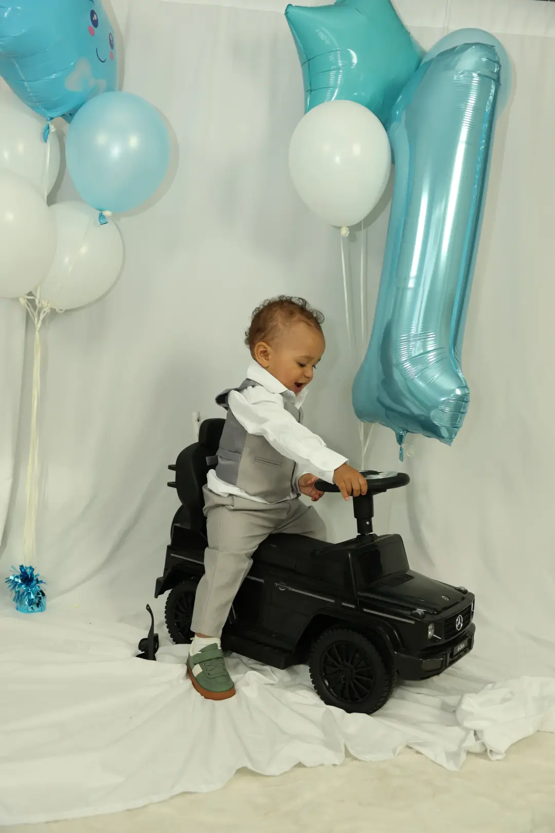 A toddler in a grey vest leans over the steering wheel of a black toy ride-on car, framed by blue and white birthday balloons.