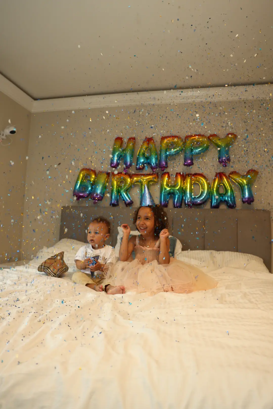 Two children celebrate on a bed showered in colorful confetti beside a rainbow HAPPY BIRTHDAY balloon banner.