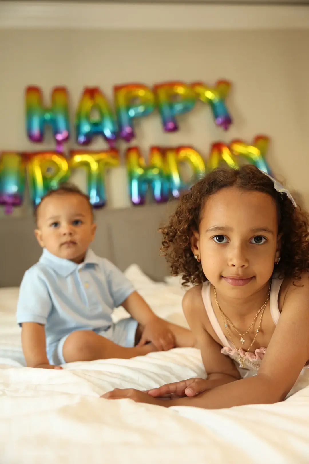 A baby boy and a young girl sit together on a bed beneath a rainbow Happy Birthday balloon banner.