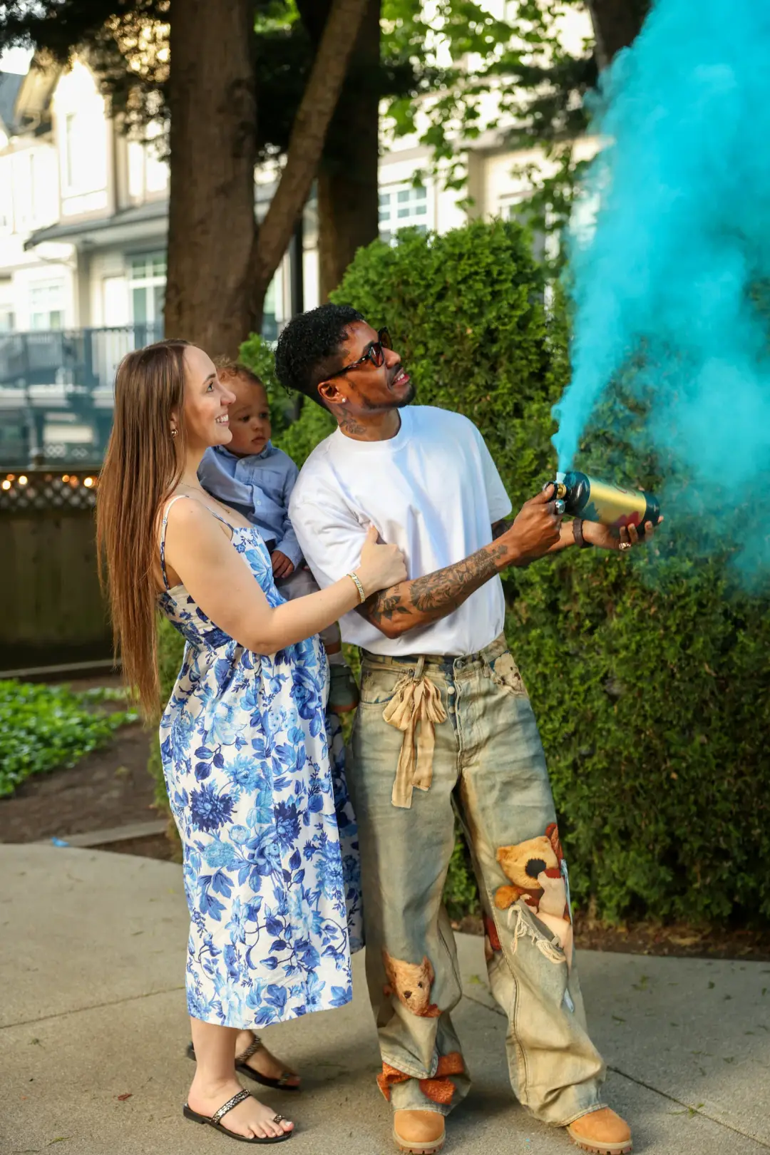 A woman holding a baby smiles as a man beside her releases teal smoke from a smoke bomb during an outdoor celebration.