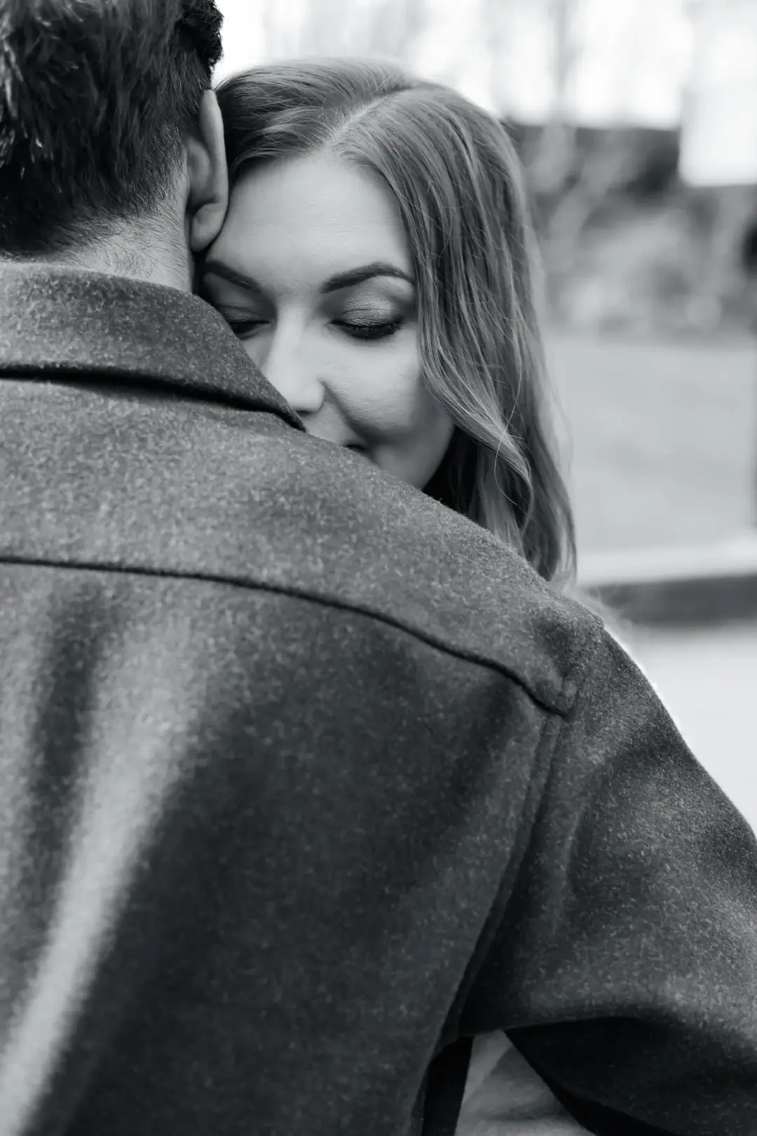 A couple in a close embrace from behind, her eyes closed against his shoulder in a soft black-and-white portrait.