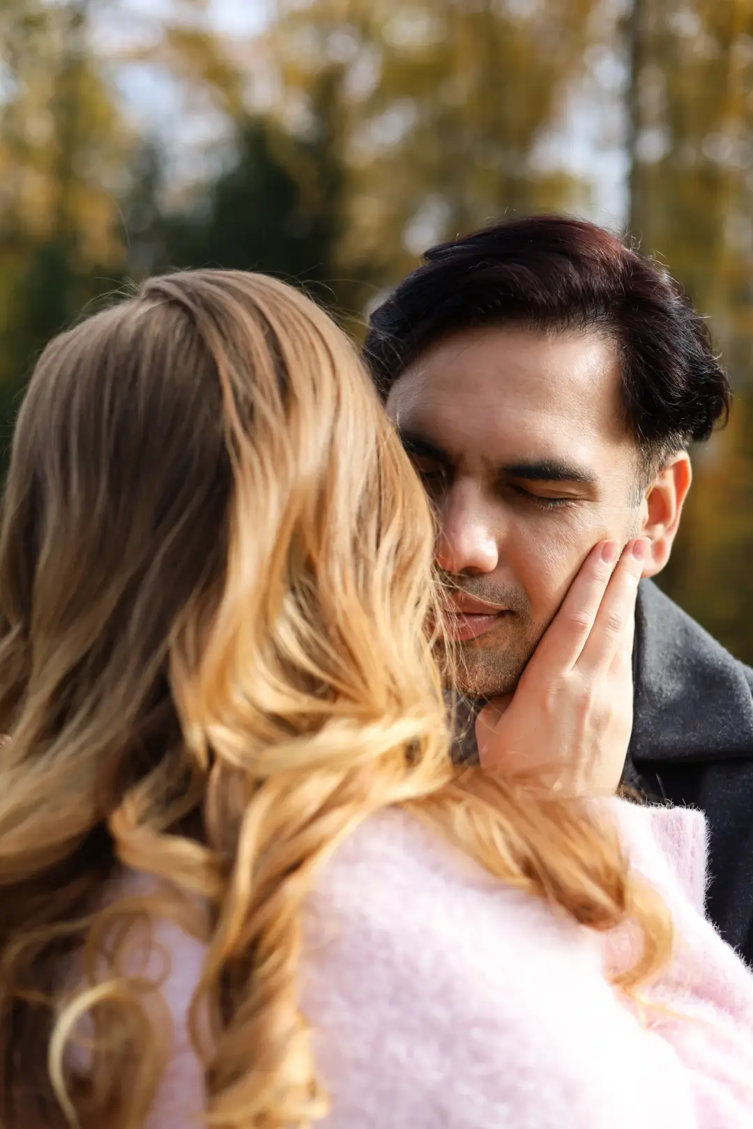 A couple standing forehead to forehead with her hand on his cheek, framed by soft autumn trees.