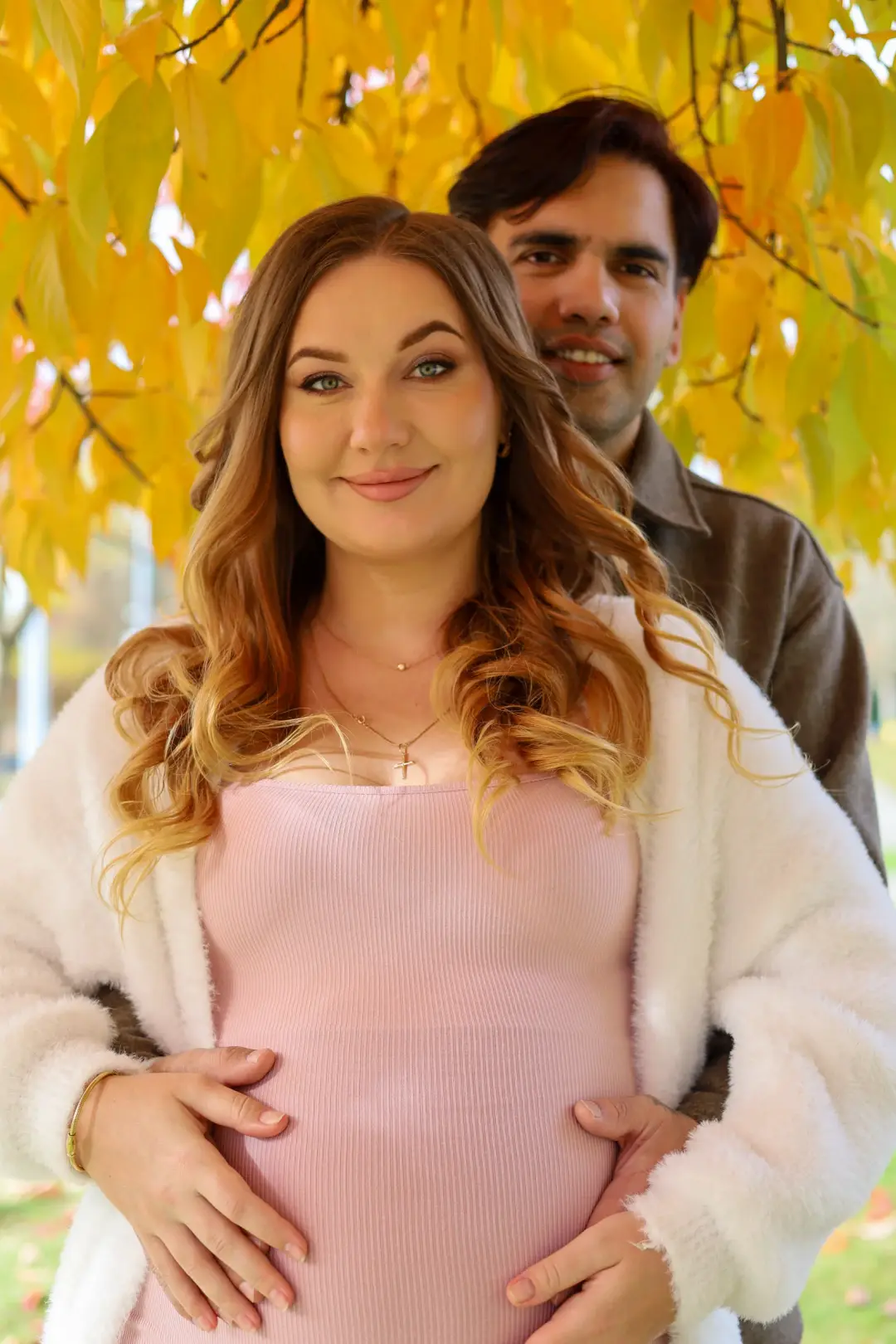 A couple smiling toward the camera under golden autumn foliage, hands resting together on her belly.