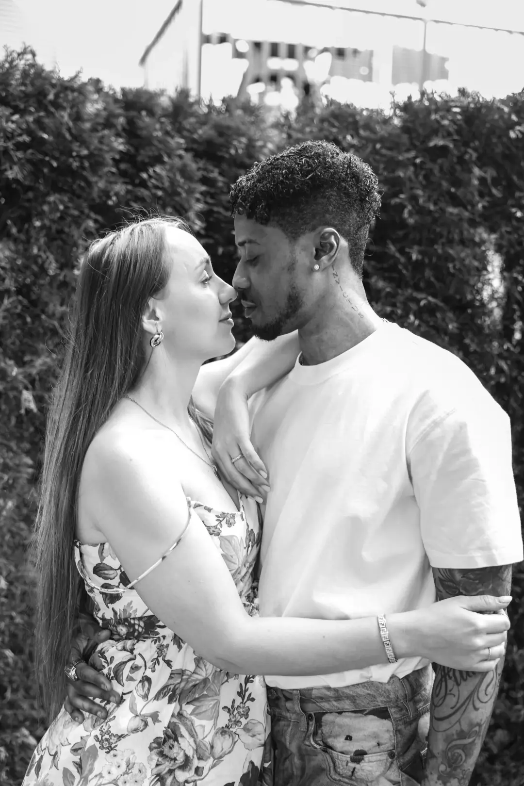 A couple standing close with noses nearly touching, her hand on his shoulder, in a black-and-white garden portrait.
