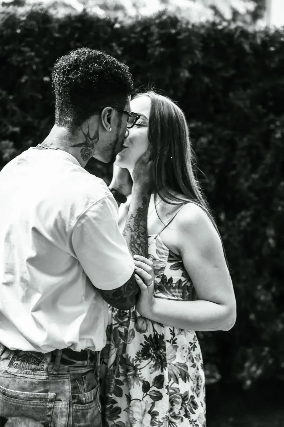 A couple sharing a tender kiss in a close black-and-white portrait against a hedge backdrop.