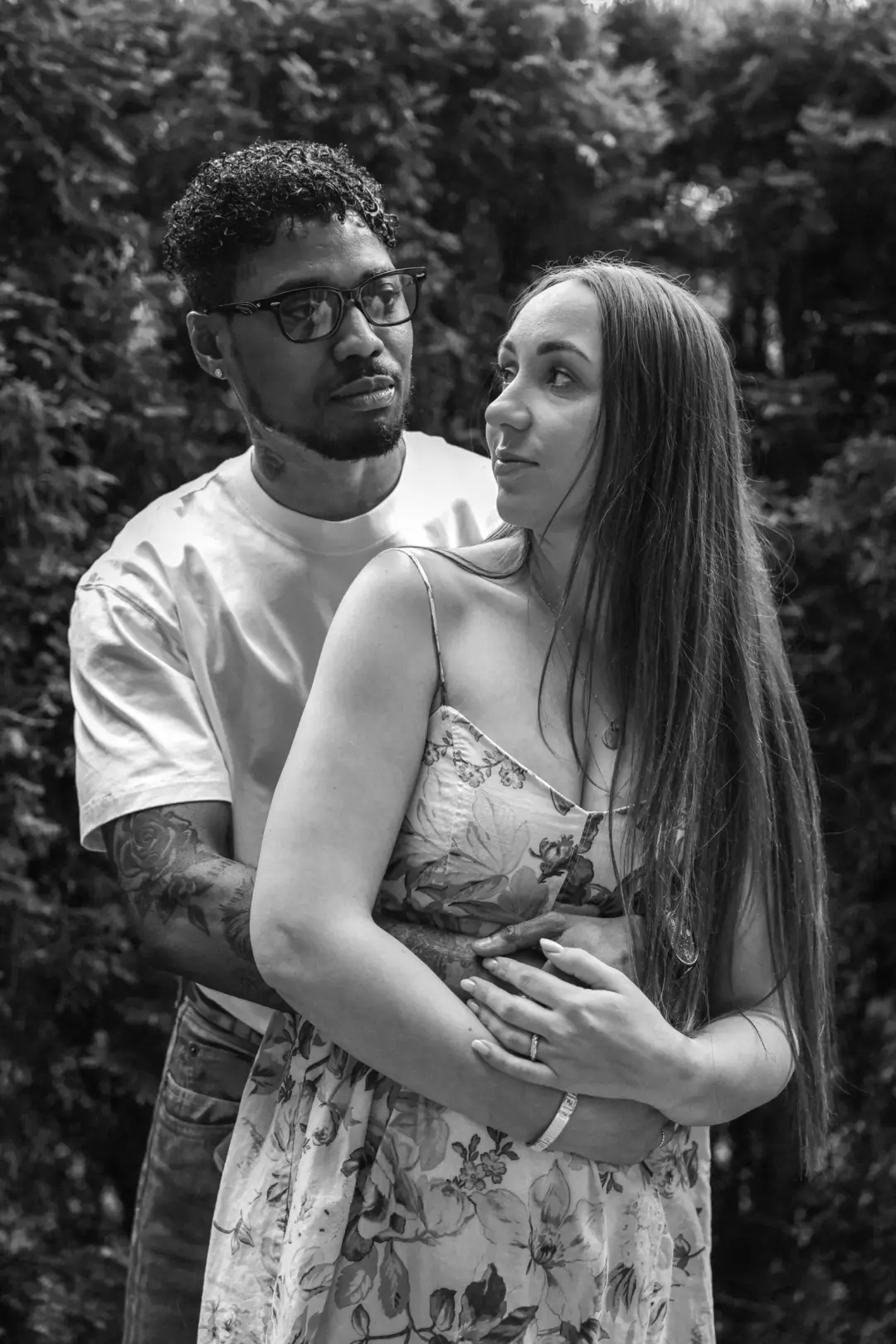 A couple embracing closely as she gazes up at him with a warm smile, set against a garden hedge in black and white.