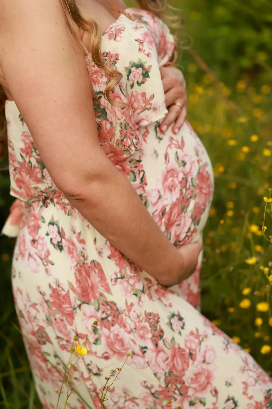 An expectant mother's hand resting on her belly in a floral dress amid wildflowers.
