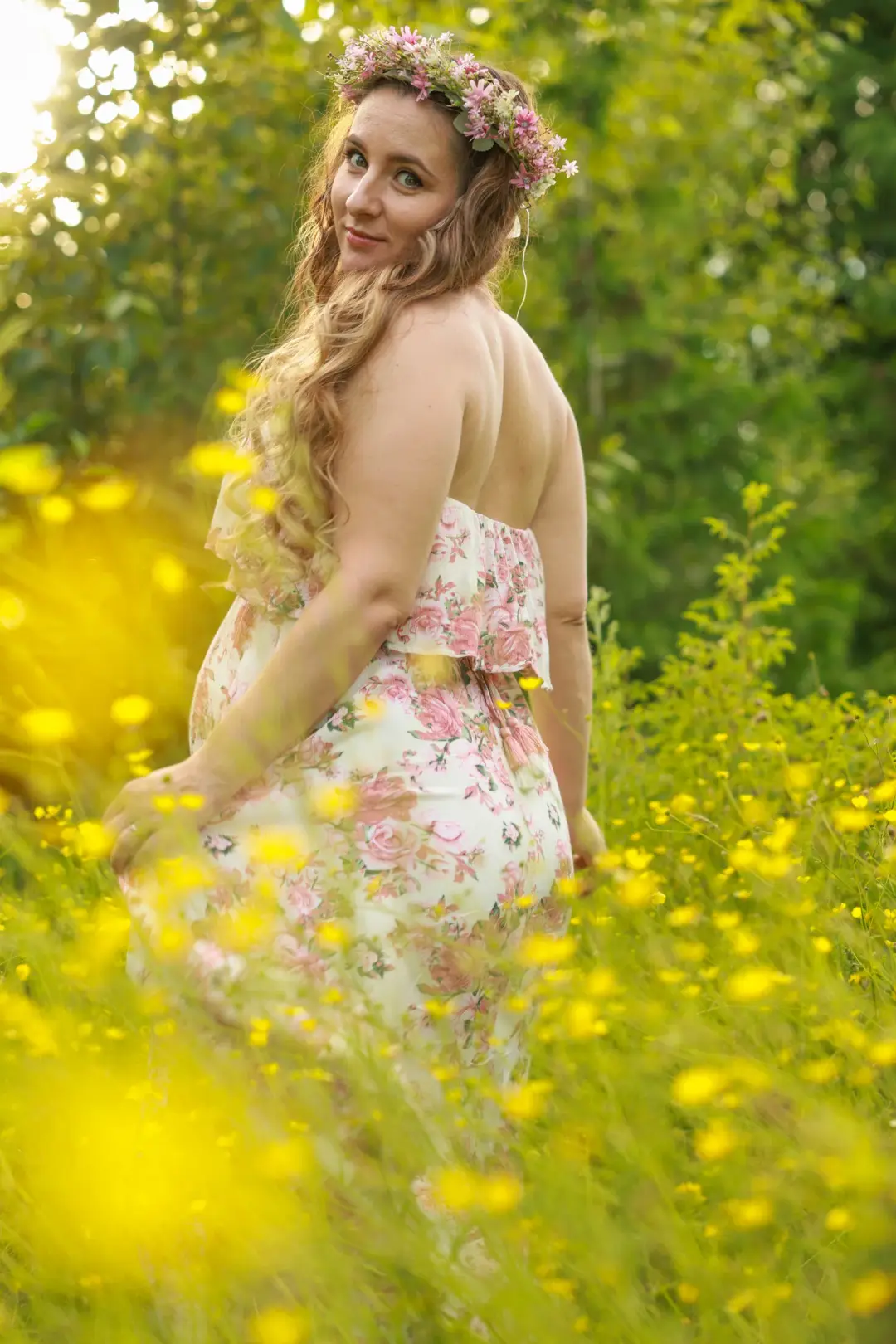 An expectant mother in a floral dress and flower crown glancing back over her shoulder in a field of yellow wildflowers.