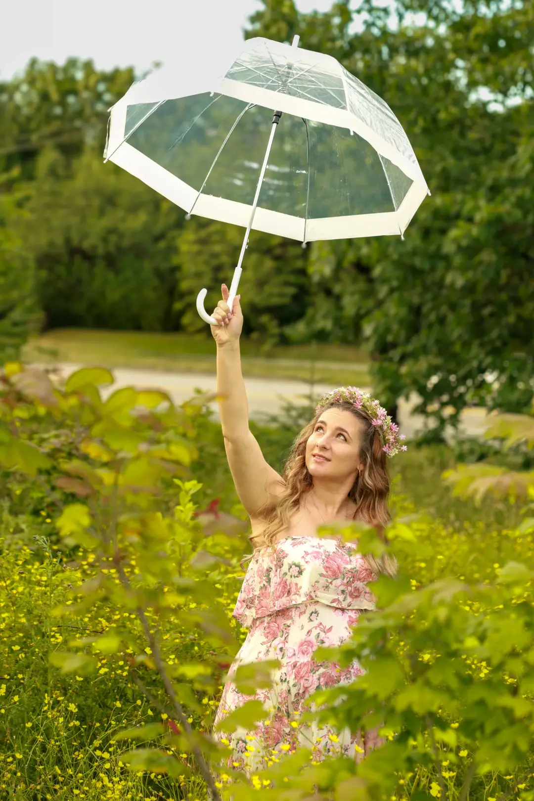 An expectant mother in a flower crown holding a clear umbrella overhead among green foliage.