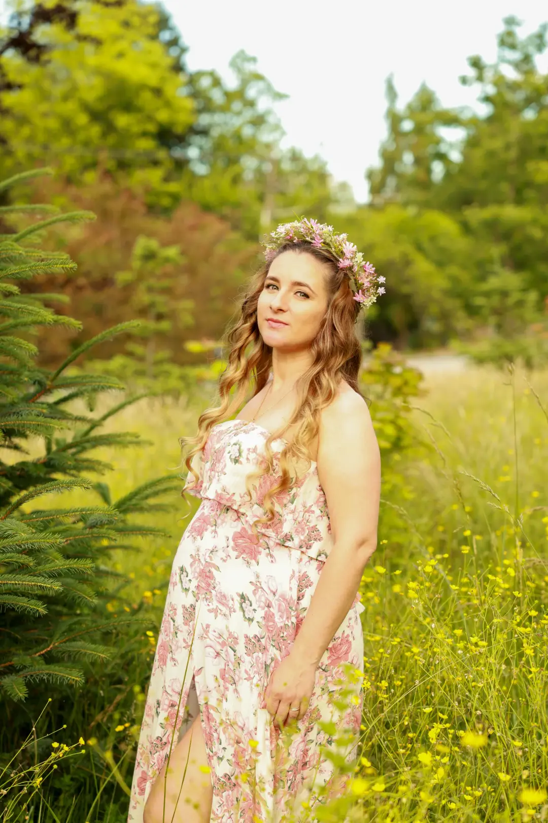 An expectant mother seen from behind in a floral dress and flower crown, standing in a field of yellow flowers.