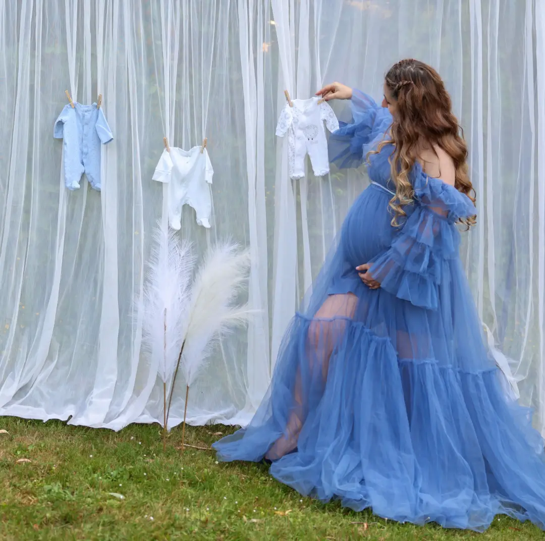 An expectant mother in a flowing blue tulle gown reaching for tiny baby outfits pinned to a sheer curtain.