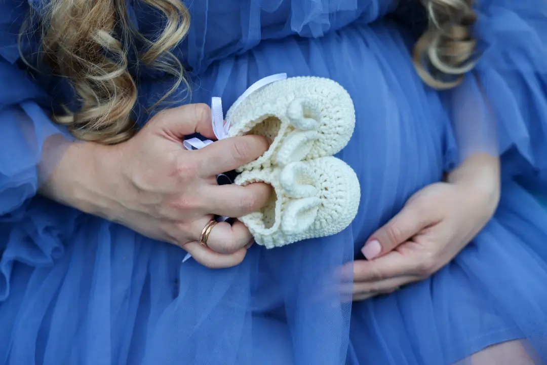 A close-up of hands holding small white knitted baby booties against a pregnant belly.
