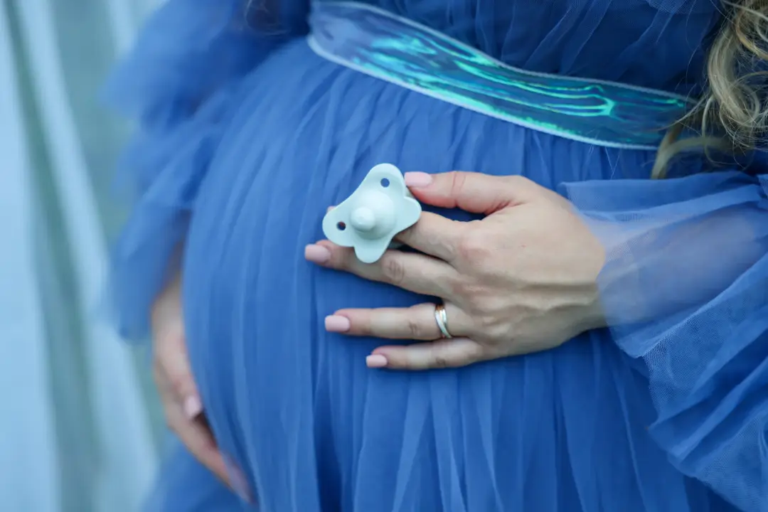 A close-up of a hand holding a baby pacifier against a pregnant belly in a blue tulle dress.
