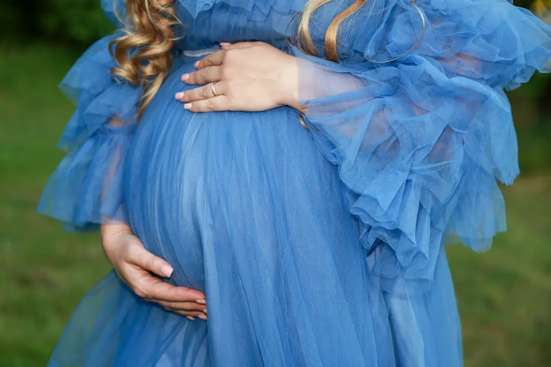 A close-up of hands cradling a pregnant belly in a ruffled blue tulle dress outdoors.