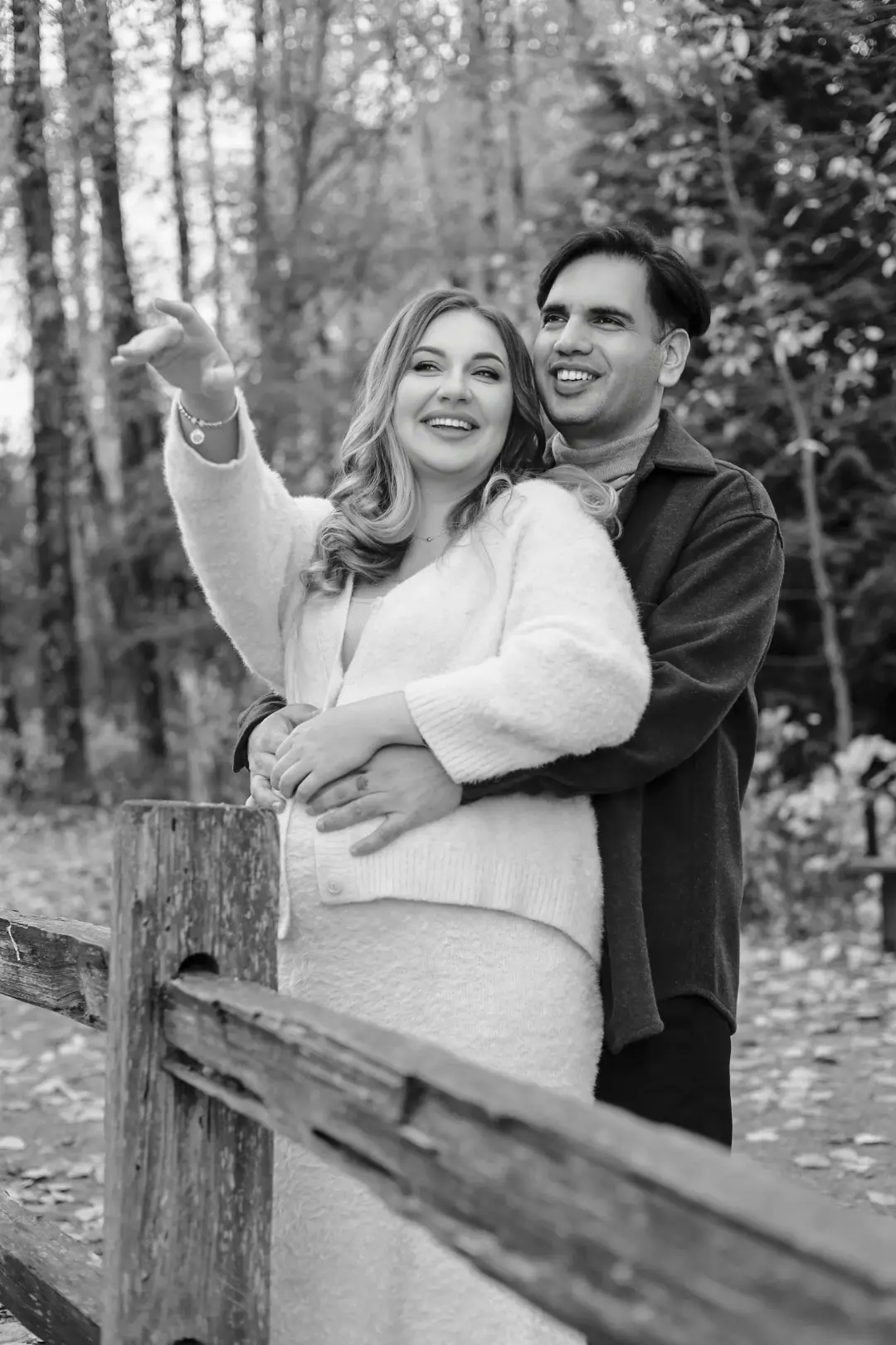 A black-and-white portrait of a couple embracing outdoors, the mother pointing and laughing as the father holds her from behind.