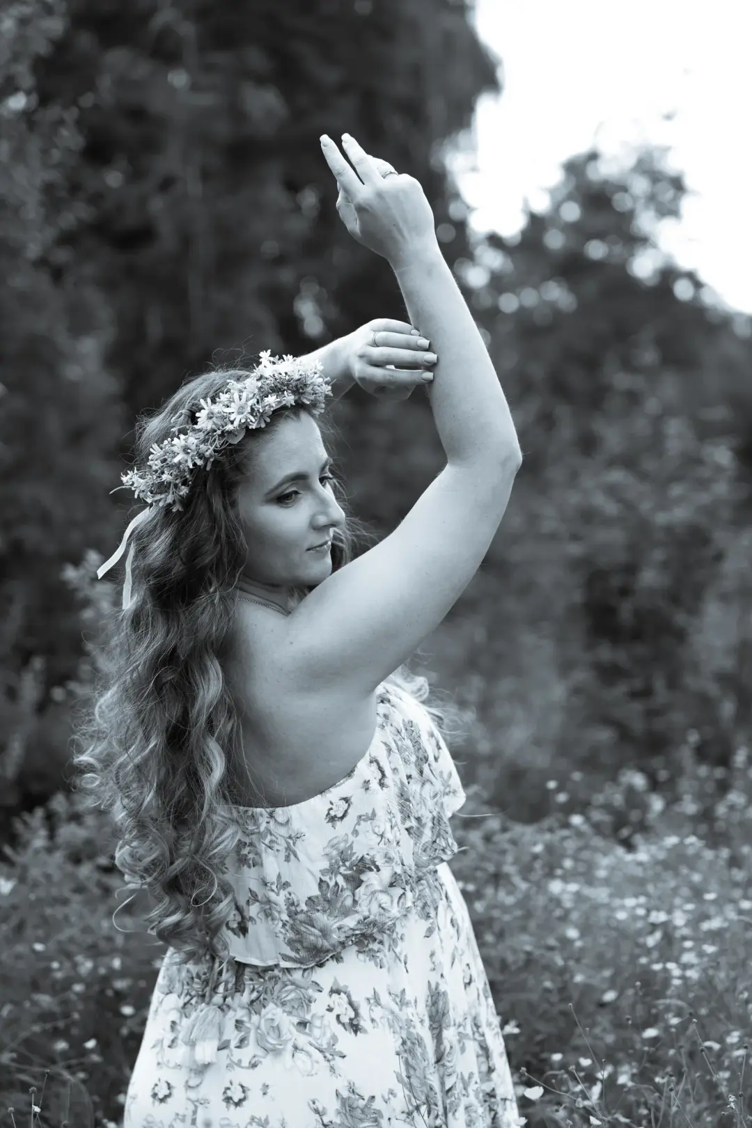 A black-and-white portrait of an expectant mother in a flower crown gracefully raising her arm in a meadow.