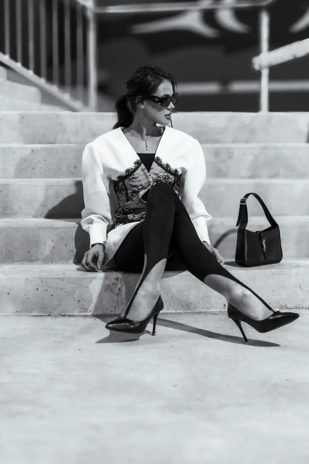 Black-and-white fashion portrait of a woman in a white blazer and lace corset seated on concrete steps, sunglasses on and a handbag beside her