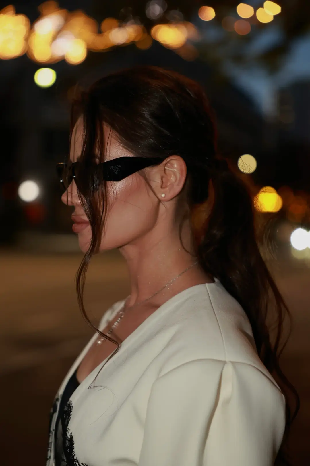 Nighttime profile portrait of a woman in sunglasses and a cream blazer, softly lit against blurred city lights
