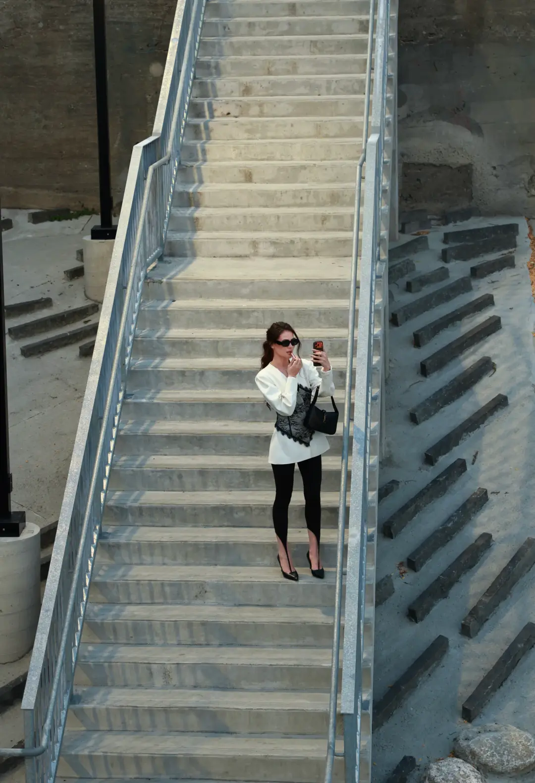 Aerial view of a woman in a white blazer and black leggings pausing on an outdoor concrete staircase to check her phone