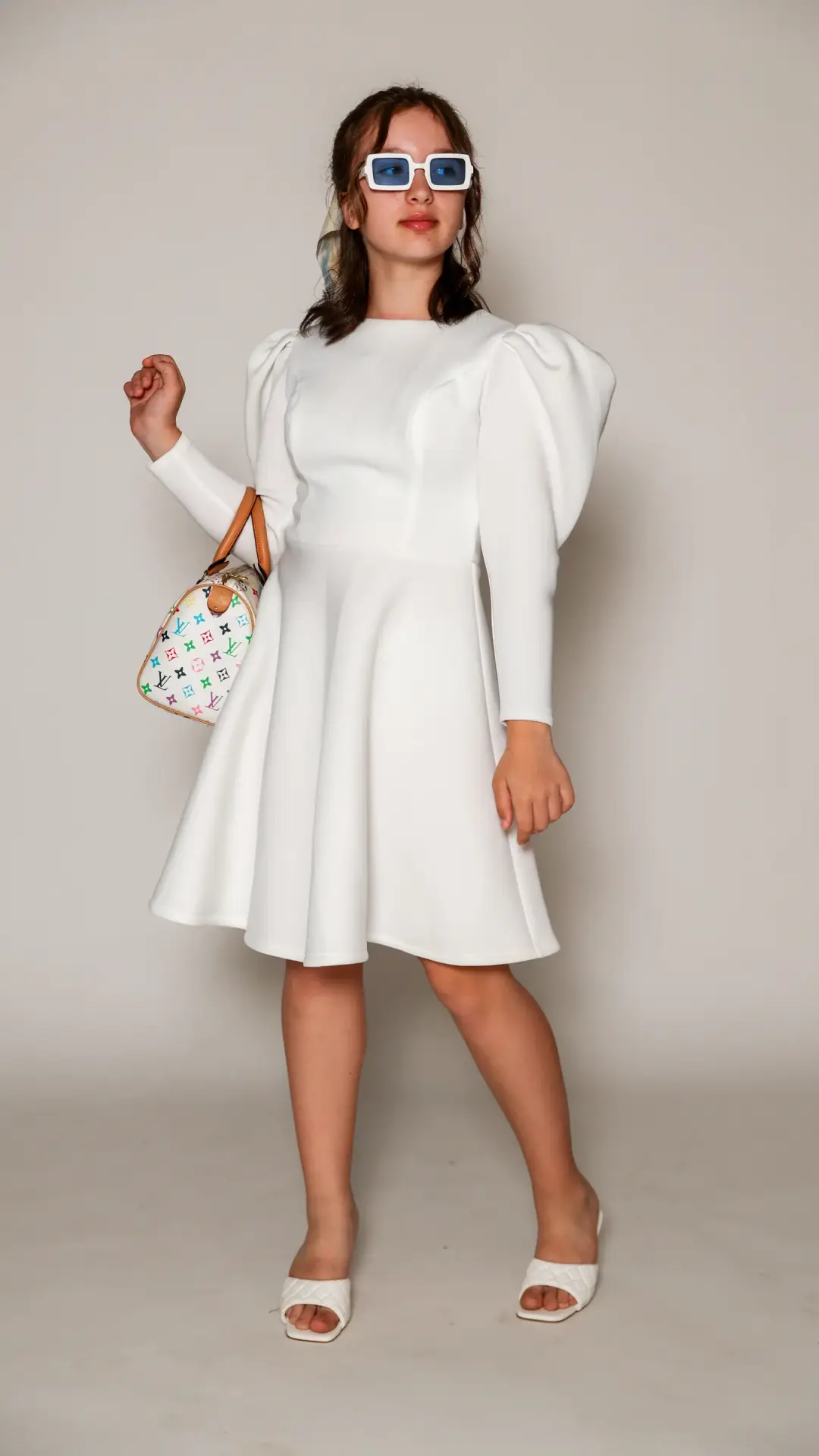 Full-length studio portrait of a young woman in a white puff-sleeve dress and blue sunglasses, holding a colorful handbag