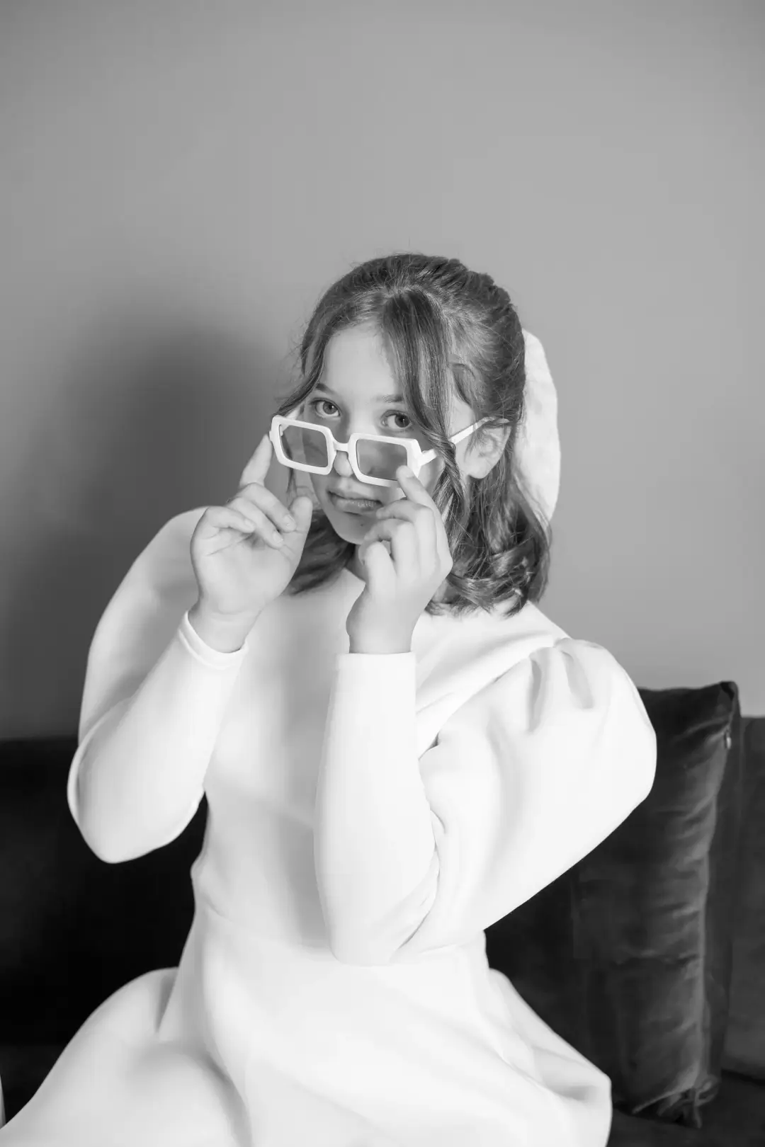 Black-and-white portrait of a young woman in a white dress peering over her sunglasses with a playful expression