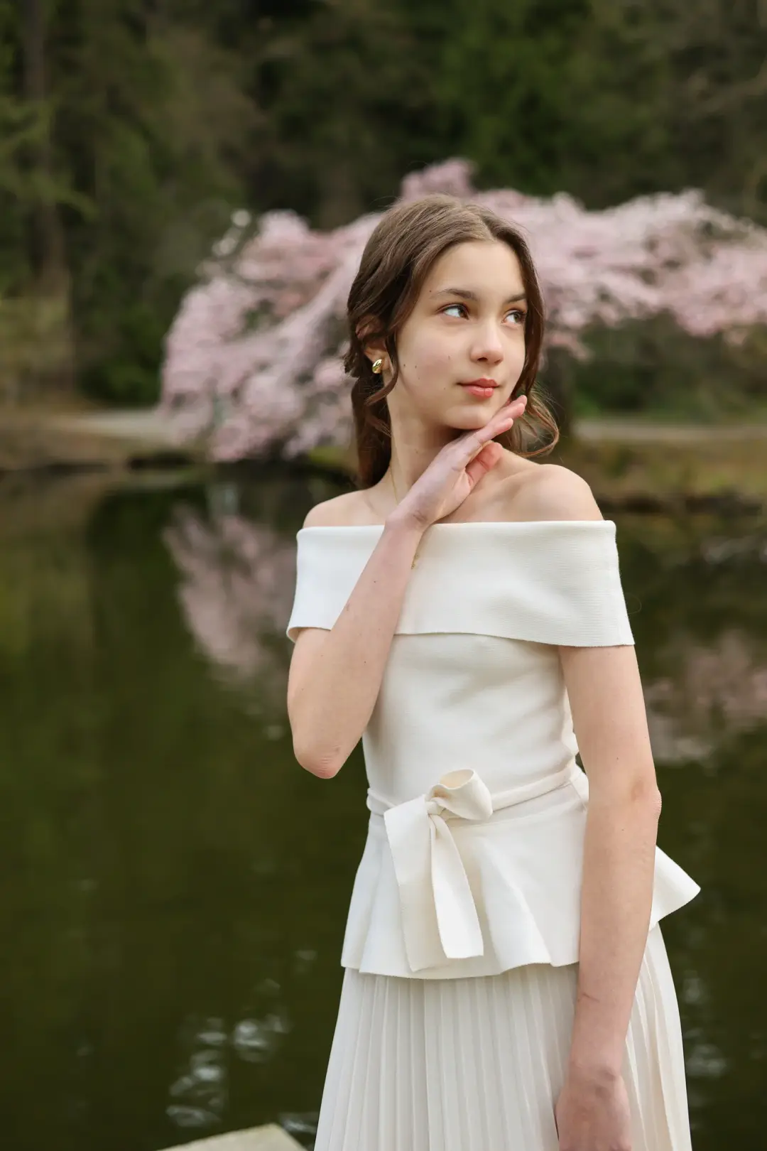 Outdoor portrait of a young woman in an off-shoulder cream top, hand at her chin, with cherry blossoms and a pond in the background
