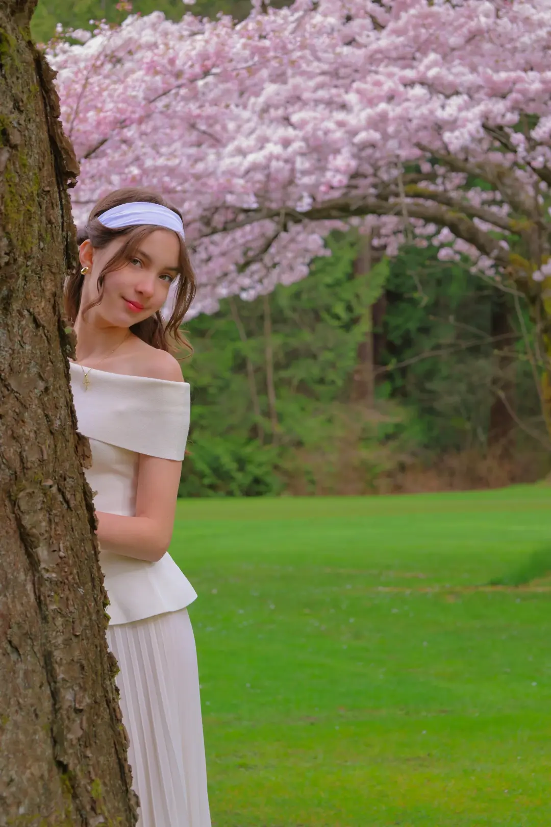Outdoor portrait of a young woman leaning against a tree trunk, cherry blossoms framing her in a sunlit park