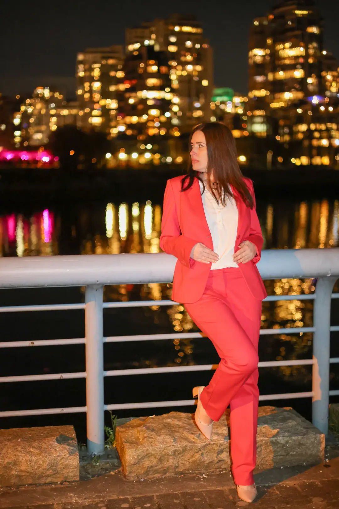 Evening portrait of a woman in a coral suit leaning against a waterfront railing, city skyline glowing behind her