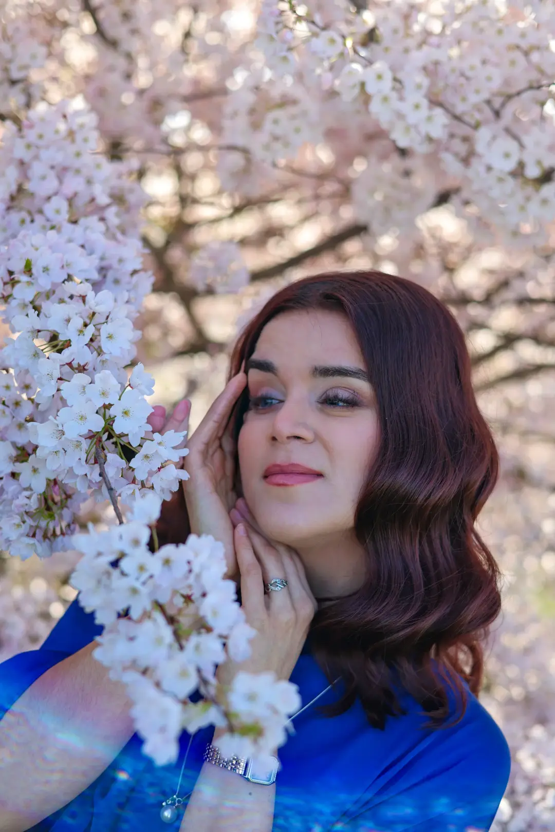 Close-up portrait of a woman in a blue dress framed by cherry blossom branches, hand resting near her face