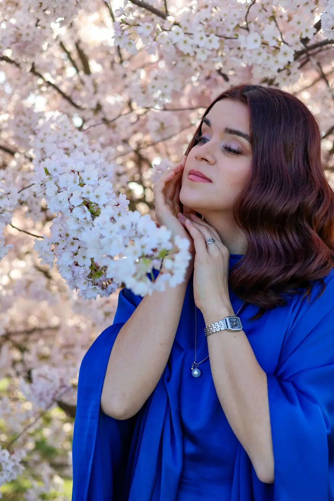 Close-up portrait of a woman in a blue dress with hands clasped near her chin, surrounded by cherry blossoms