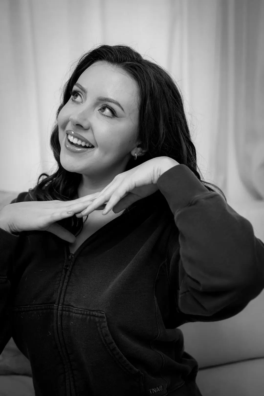 Black-and-white candid portrait of a woman laughing, hand raised near her shoulder, against a soft curtain backdrop