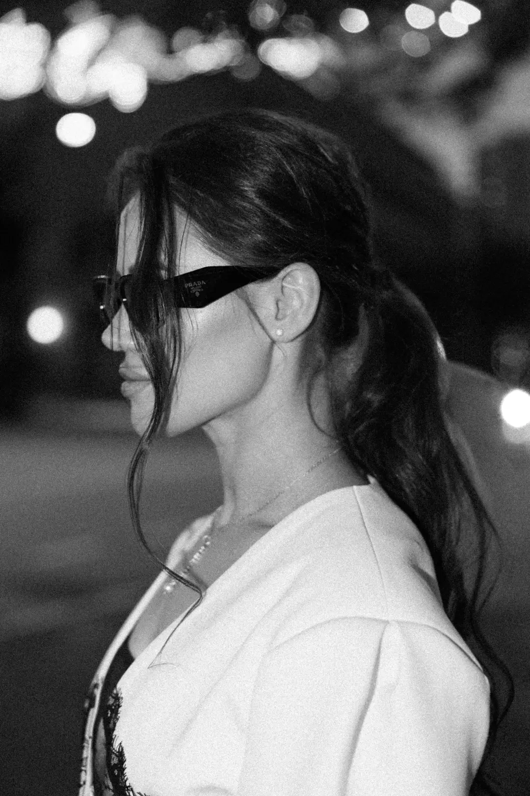 Black-and-white nighttime profile portrait of a woman in sunglasses and a white blazer, city lights blurred behind her