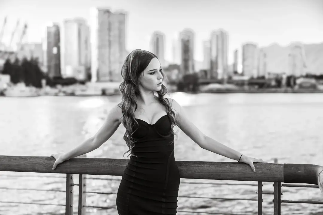 Black-and-white waterfront portrait of a woman in a black dress leaning on a wooden railing, city skyline across the water